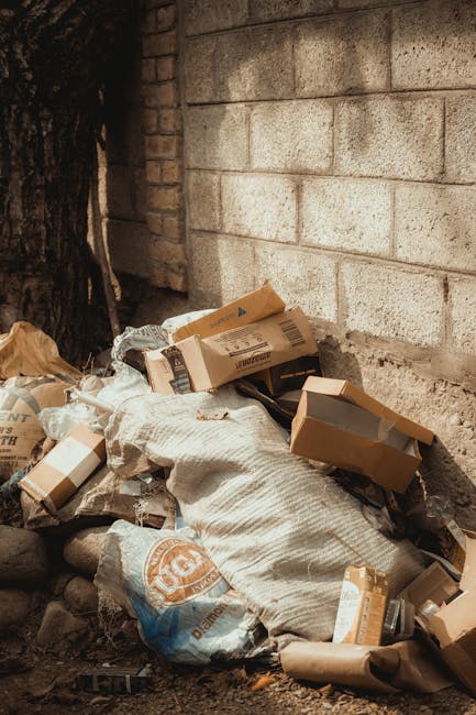 A person wearing a yellow and blue checkered shirt, blue jeans, and bright green gloves is holding open a large black rubbish bag, which appears to be made of thick plastic with a shiny finish. The in
