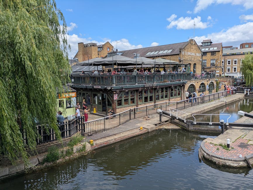 A multi-level brick building with a dark timber structure housing a restaurant or pub, featuring large windows on the ground floor and an outdoor upper terrace with a canopy covering, occupied by nume